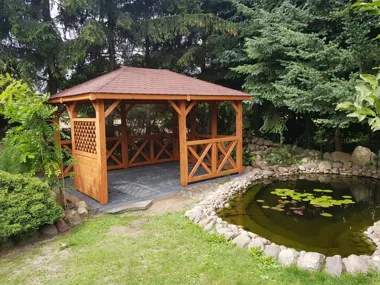 Drewniana altana przy oczku wodnym z liliami, otoczona drzewami i krzewami.