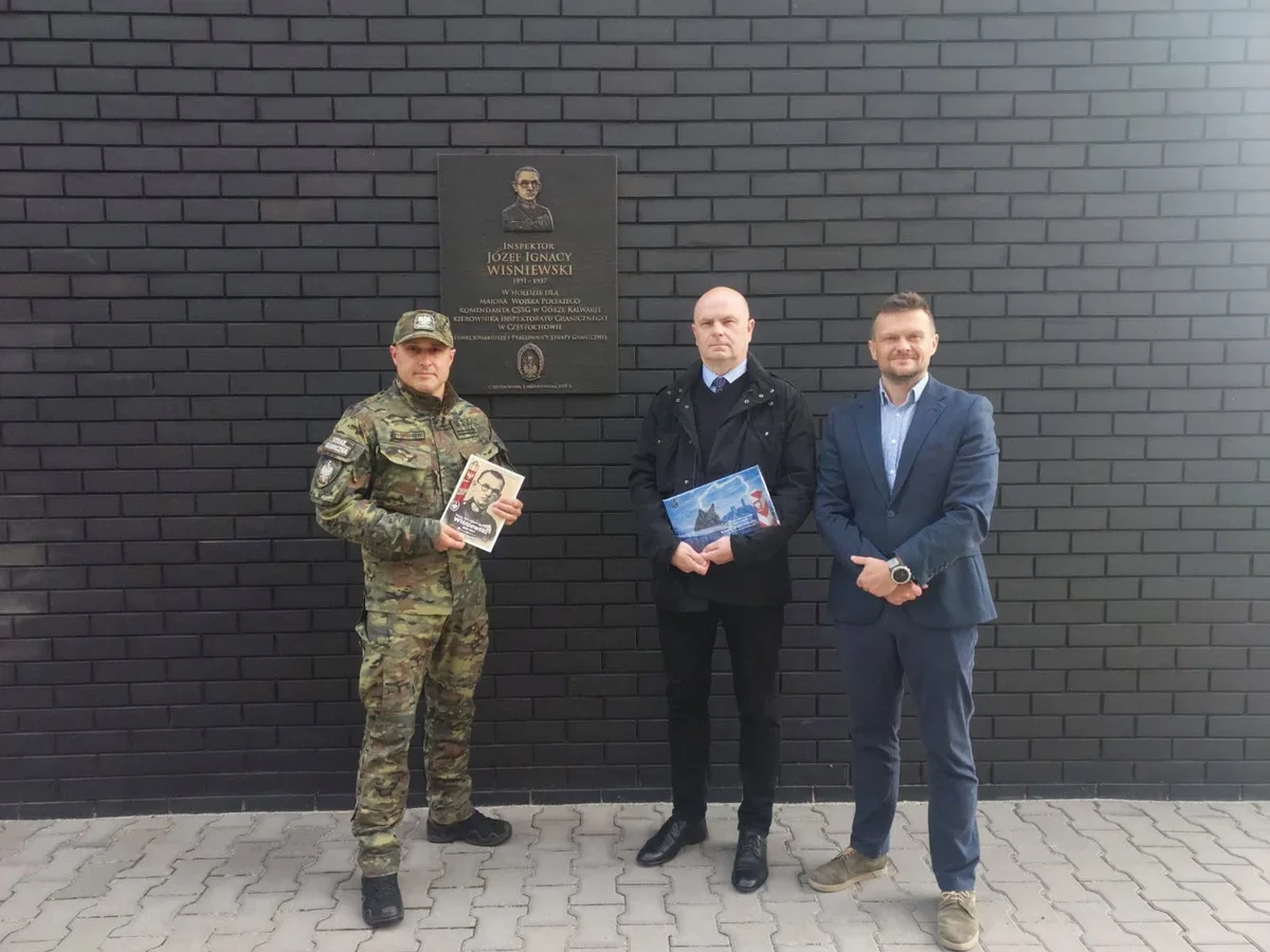 Trzech mężczyzn stoi przed czarną ceglaną ścianą z tablicą pamiątkową; żołnierz w mundurze i dwaj mężczyźni w garniturach trzymają broszury.