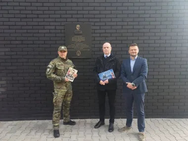 Trzech mężczyzn stoi przed czarną ceglaną ścianą z tablicą pamiątkową; żołnierz w mundurze i dwaj mężczyźni w garniturach trzymają broszury.
