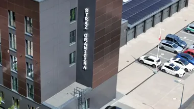 Budynek placówki Straży Granicznej z widocznym neonem na elewacji oraz zaparkowanymi samochodami służbowymi na parkingu.