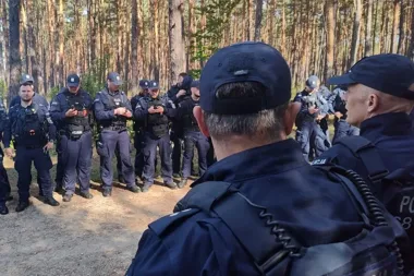 Grupa policjantów w mundurach i czapkach stoi w lesie podczas odprawy lub briefing.