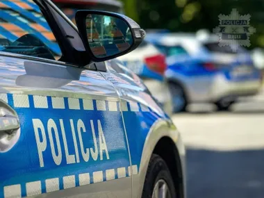 Fragment policyjnego radiowozu z napisem „Policja” zaparkowanego na ulicy, w tle rozmyte inne pojazdy.