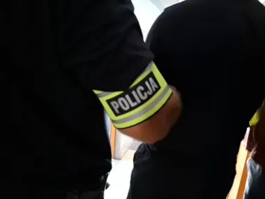 Funkcjonariusz policji z opaską oznaczoną napisem POLICJA na ramieniu, widziany z tyłu w pomieszczeniu.