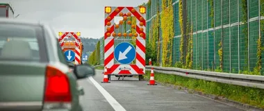 Samochód osobowy jedzie za dwoma pojazdami oznakowania drogowego wskazującymi kierunek jazdy na wielopasmowej drodze obok ekranów dźwiękochłonnych porośniętych