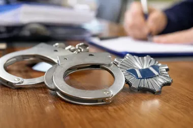 Kajdanki i policyjna odznaka leżą na drewnianym biurku, w tle rozmyte dłonie osoby piszącej w notatniku.