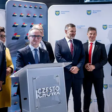 Grupa osób w strojach formalnych stoi przy mównicy z napisem Częstochowa na tle ścianek z herbem Województwa Śląskiego.