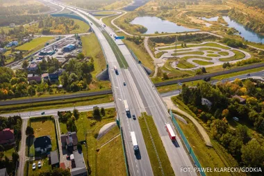 Wielopasmowa autostrada biegnie przez zielony, lekko pagórkowaty teren z zabudową, torami kolejowymi i stawami w tle.