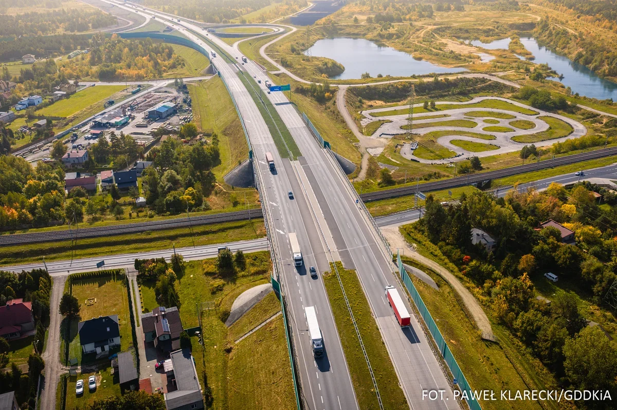 Wielopasmowa autostrada biegnie przez zielony, lekko pagórkowaty teren z zabudową, torami kolejowymi i stawami w tle.